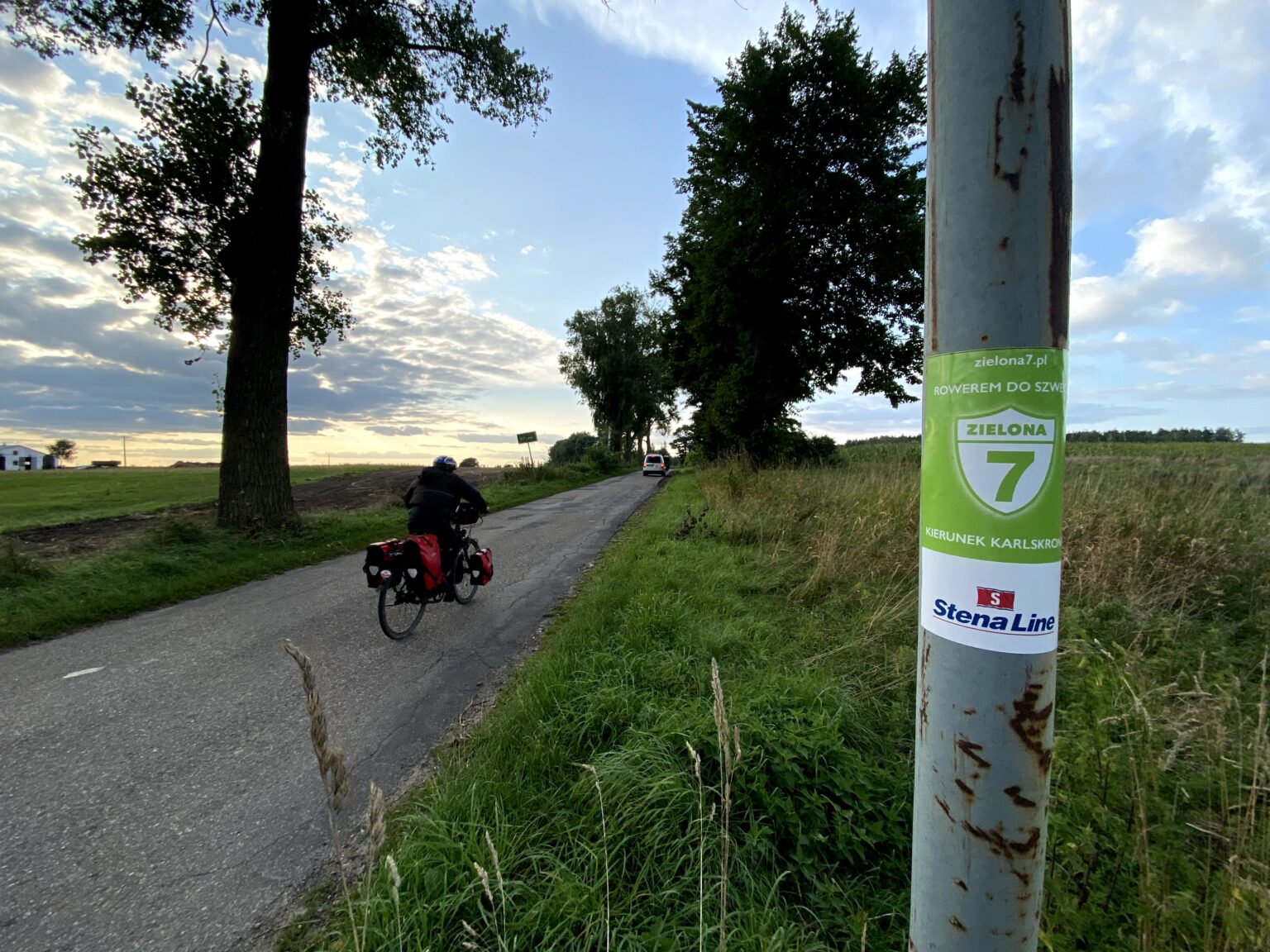 Rowerzysta z sakwami na asfaltowej drodze wśród pól, na słupie naklejka Zielonej 7
